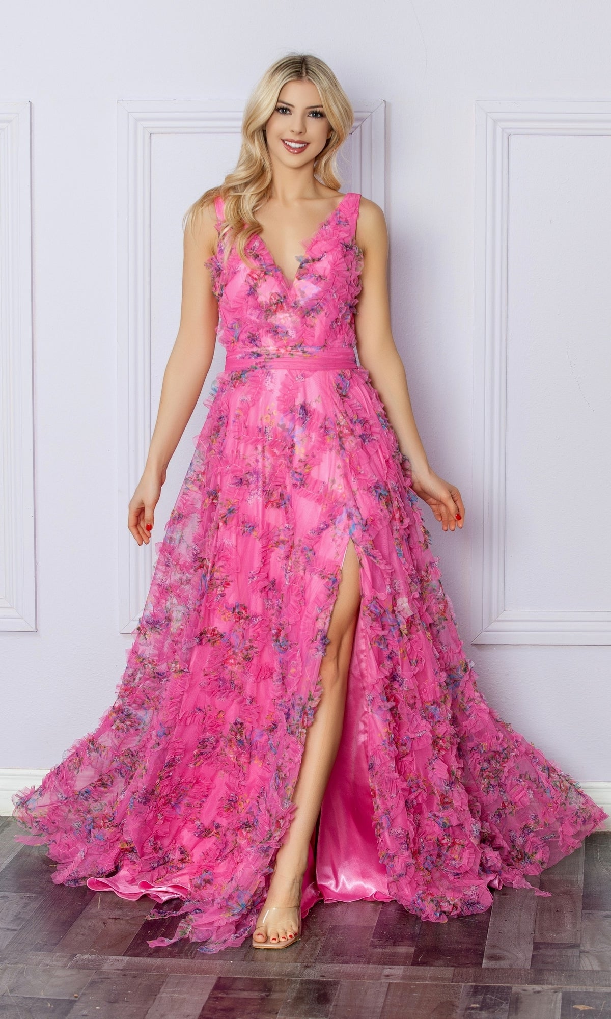 A woman stands indoors smiling at the camera, wearing the Pastel Print Long Prom Dress: Nox Anabel E1445—a floor-length pink floral gown with a deep V-neck, high slit, and sleeveless design.